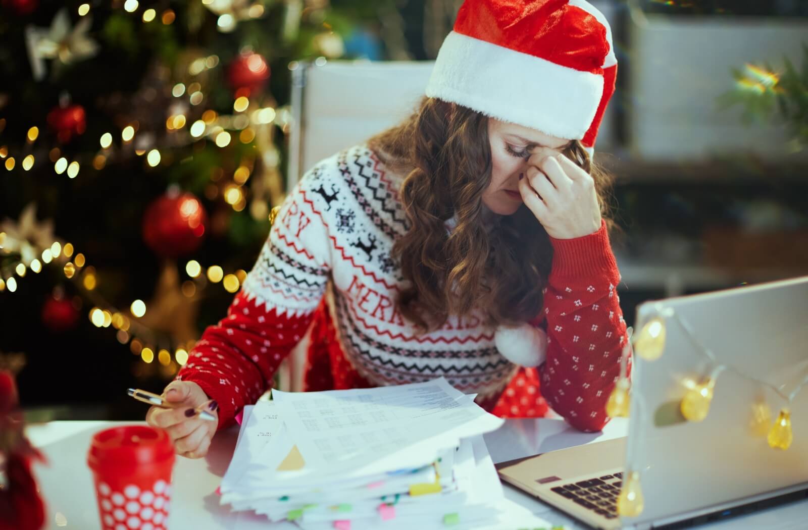 A woman wearing a Christmas sweater sits at a desk with a pen in hand, looking stressed, while a decorated Christmas tree is visible in the background.